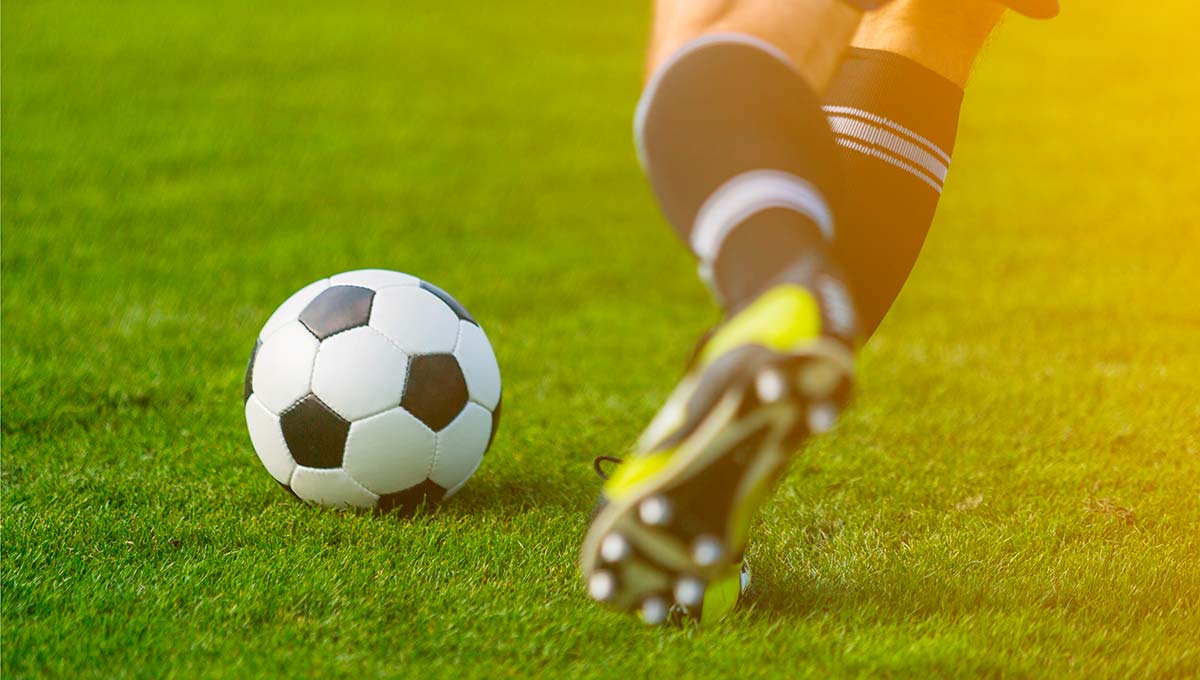 Fußballer mit schwarz-weißen Stutzen holt auf grünem Rasen mit dem linken Bein zu einem Schuss auf den vor ihm liegenden Ball aus.
